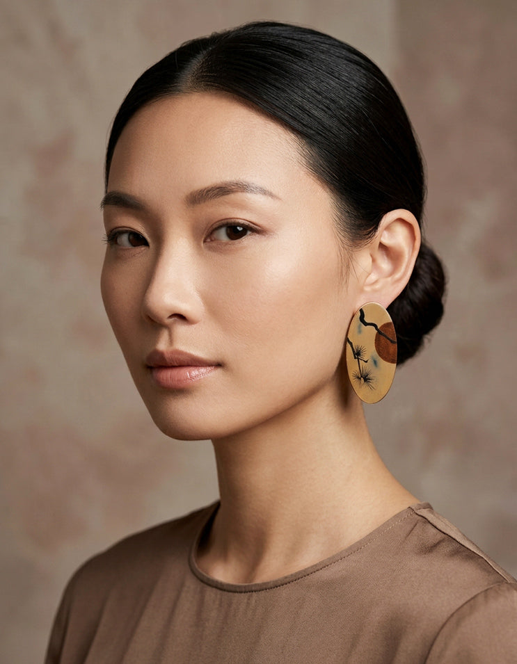 A woman wearing a miniature classical landscape Chinese lacquer wood earrings feature hand-painted pine & sunset motifs on burnished bronze lacquer. Da Qi art.